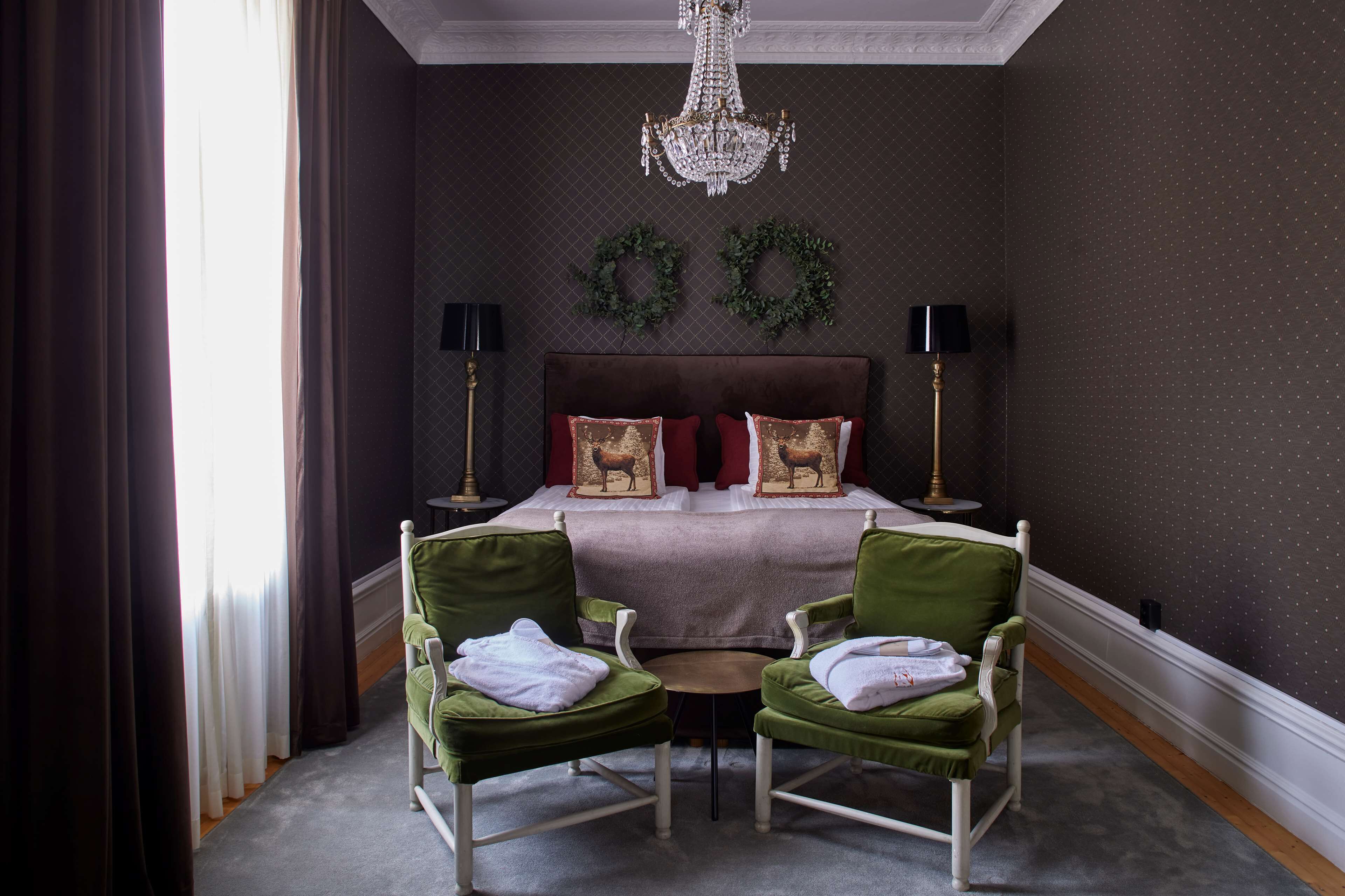 A bedroom with a large bed, two green chairs, a chandelier, and a small table.