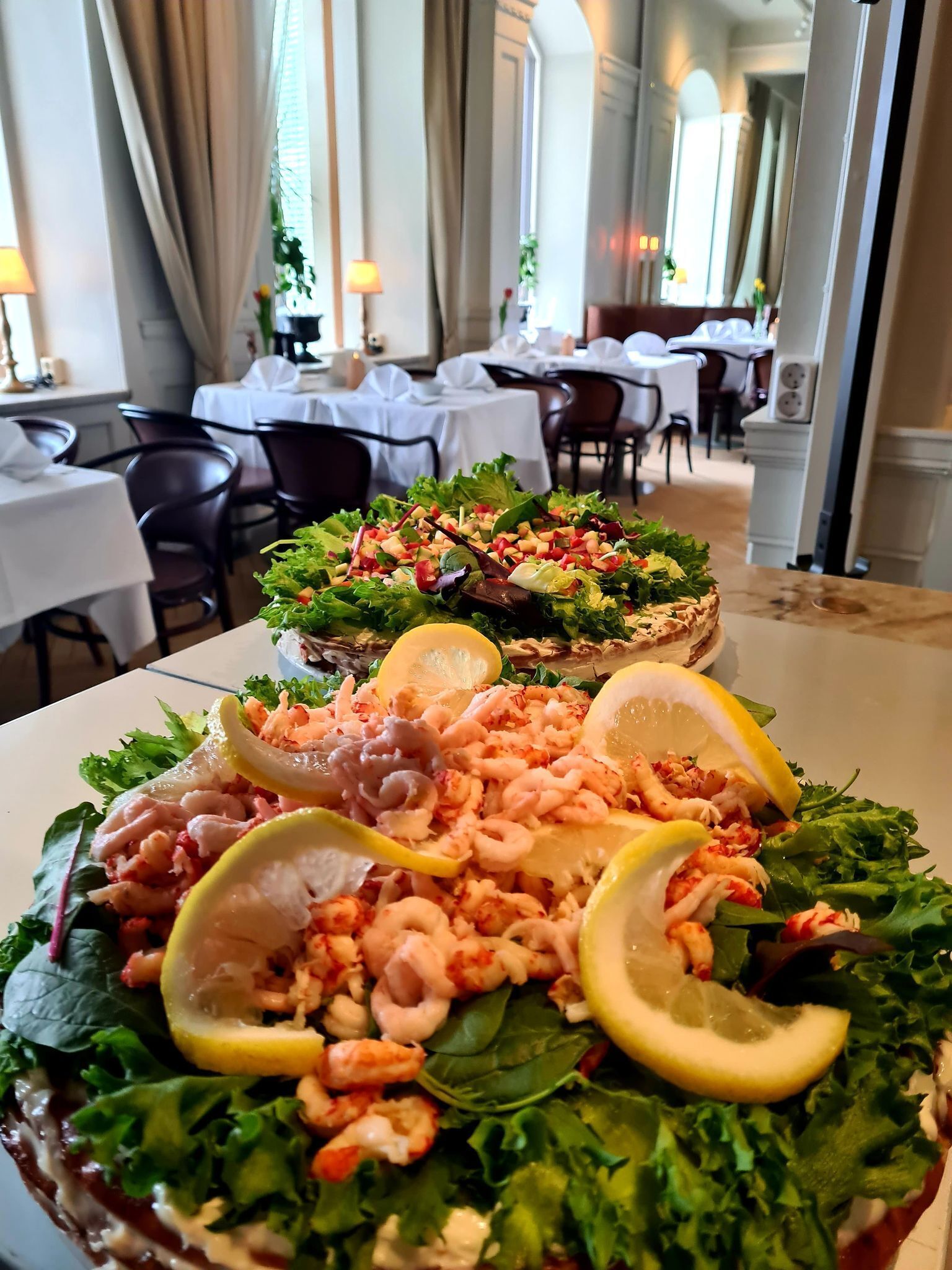 A restaurant with tables set for dining, a salad with shrimp and lemon slices, and a view of the dining area.