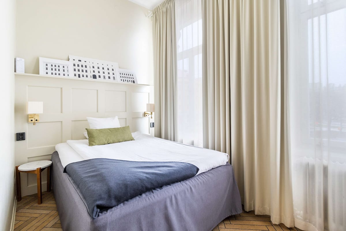 A hotel room with a large bed, a small bedside table, a nightstand with a lamp, and a window with sheer curtains.