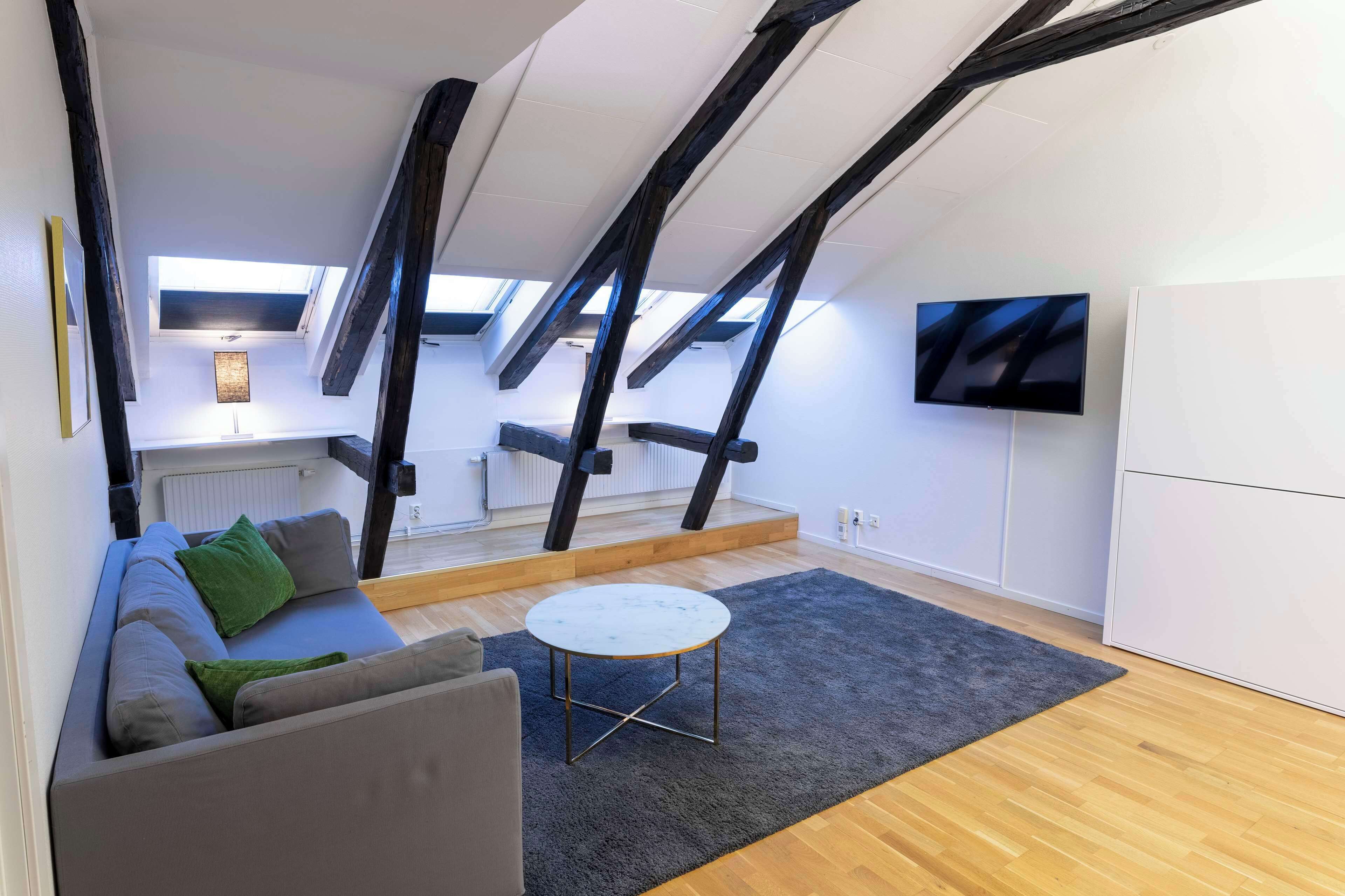 En modern lägenhet i loftstil med högt i tak, synliga takbjälkar, en soffa, ett litet runt bord och en tv som är monterad på väggen.