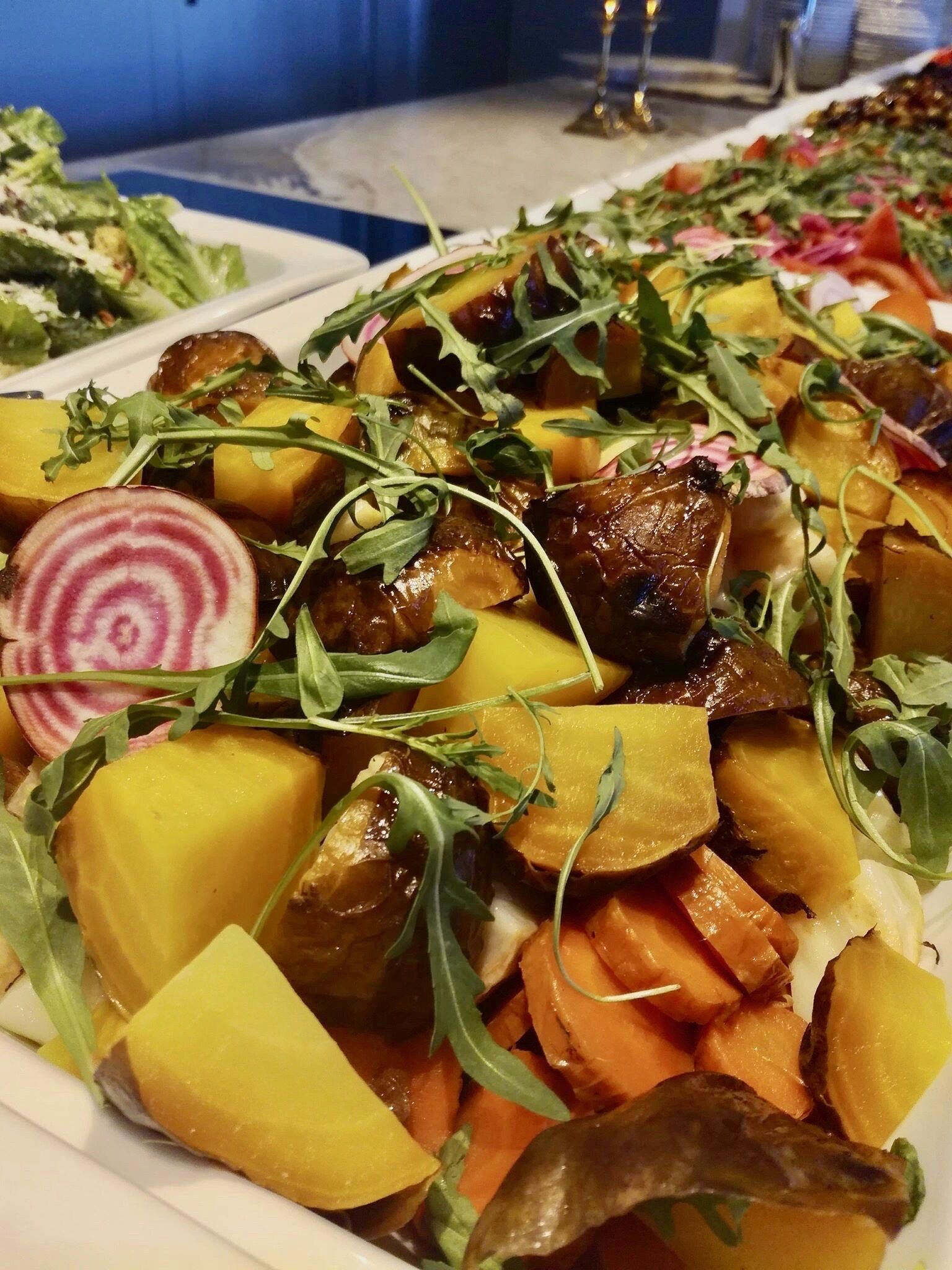 Una mesa de buffet con diversos platos, incluyendo una ensalada con rúcula y rodajas de remolacha, y un plato con calabacín y champiñones.