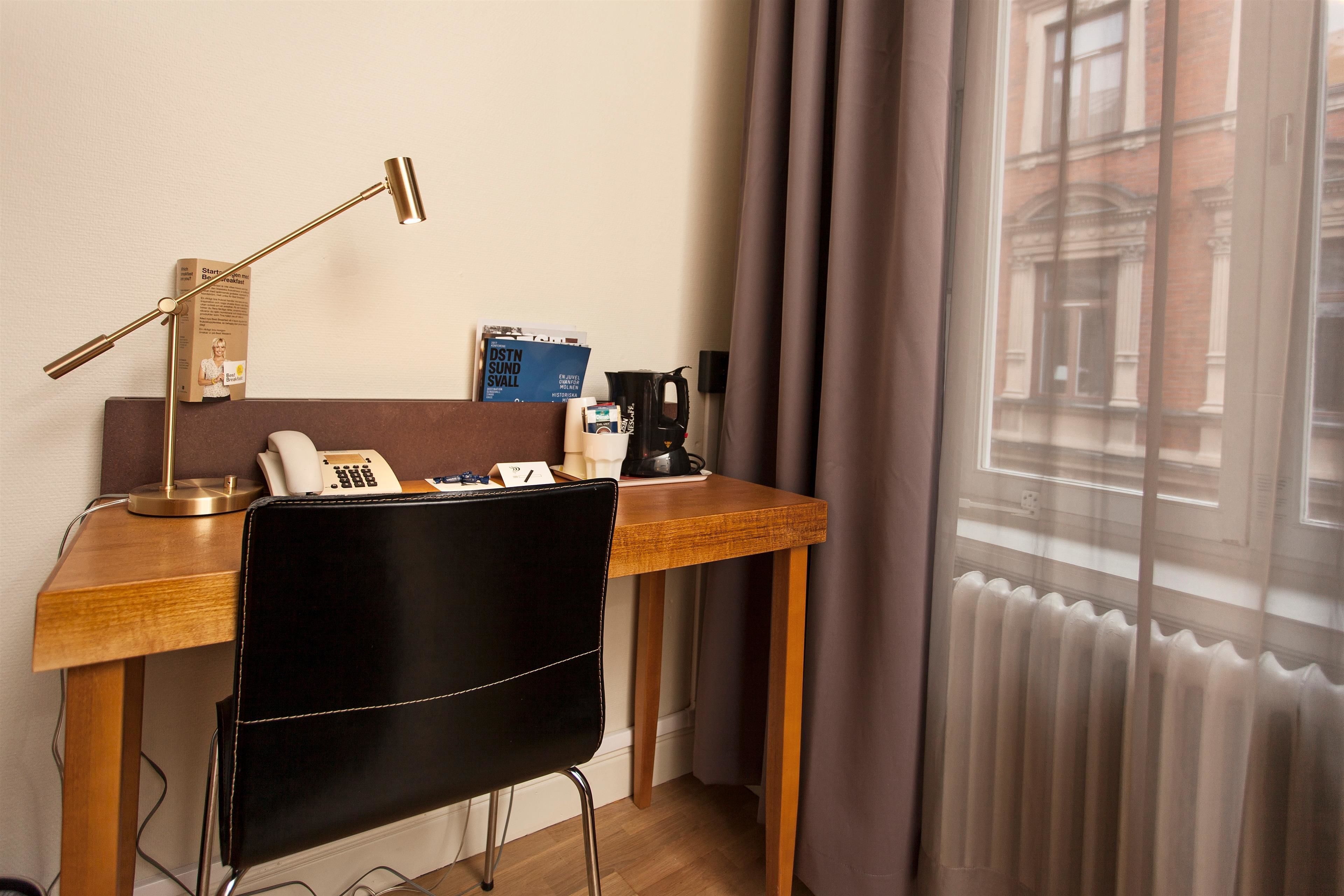 Habitación de hotel con escritorio de madera, una silla negra, teléfono, lámpara, revista y cafetera.