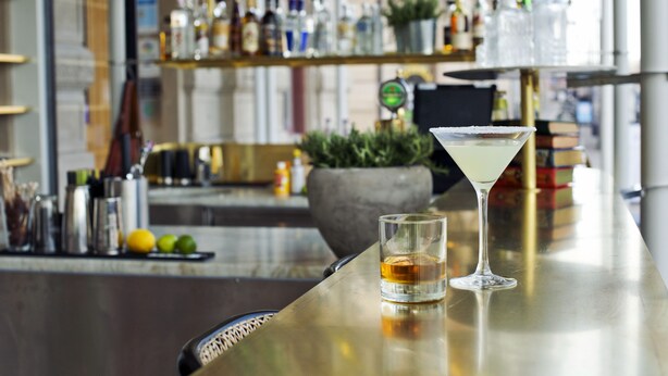 A bar area with a martini glass and a shot glass on a counter, shelves with bottles and a potted plant in the background. - Best Western Plus Hotel Baltic (Sundsvall)