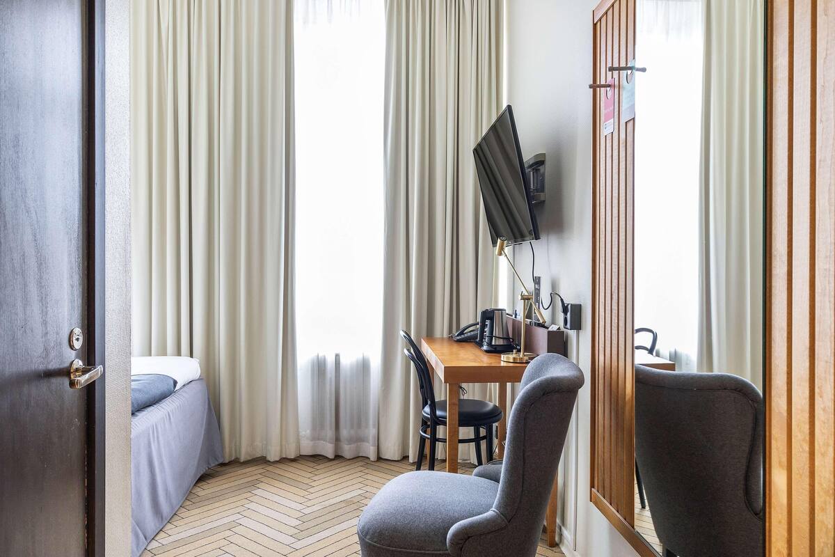 A hotel room with a bed, a desk with a chair, a TV, and a wooden door.