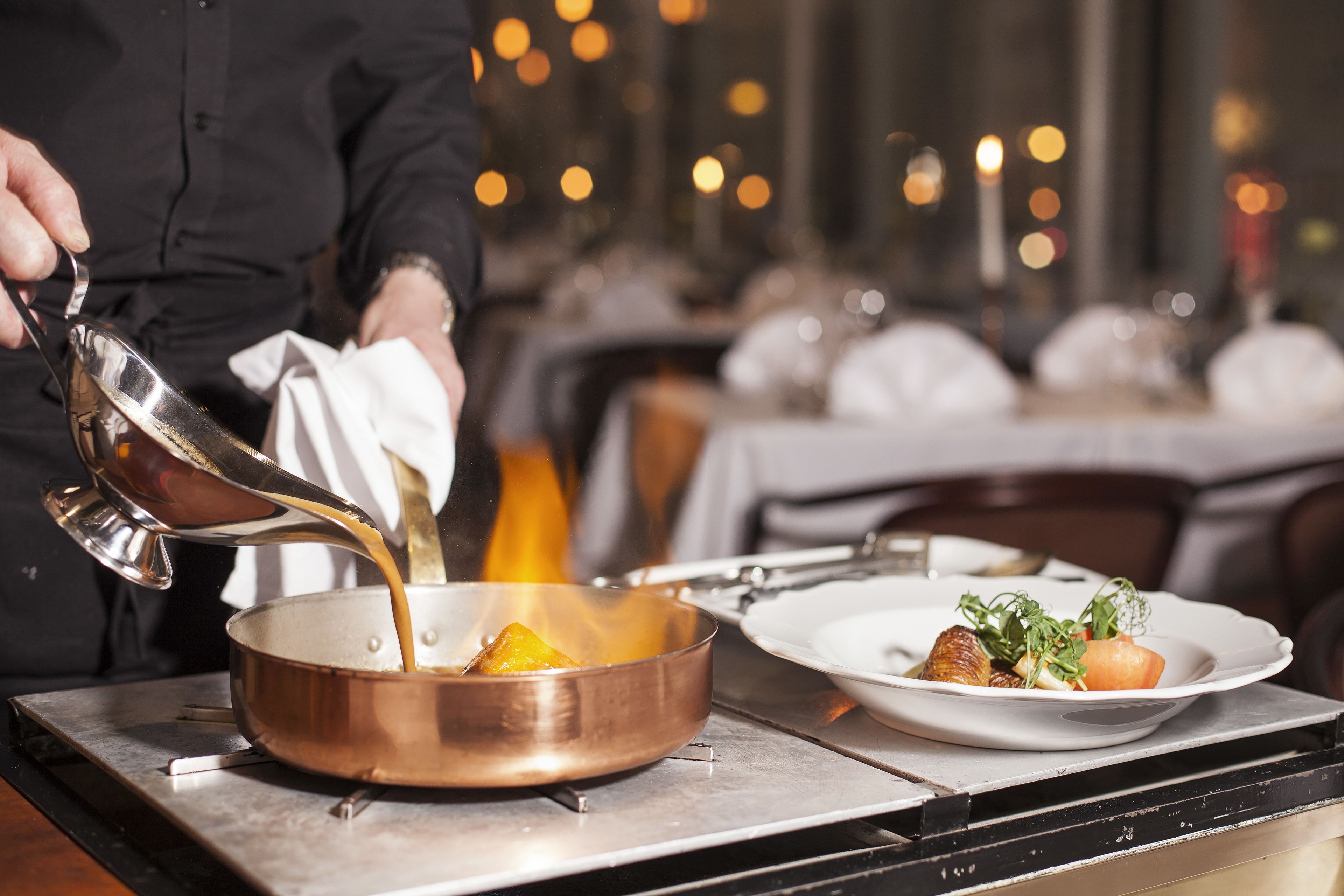 A chef pouring a liquid into a copper pan on a stove, with plates of food in the background.