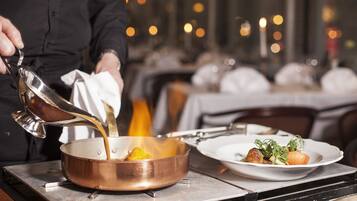 A chef pouring a liquid into a copper pan on a stove, with plates of food in the background.