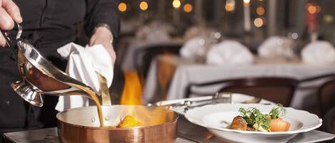 A chef pouring a liquid into a copper pan on a stove, with plates of food in the background.