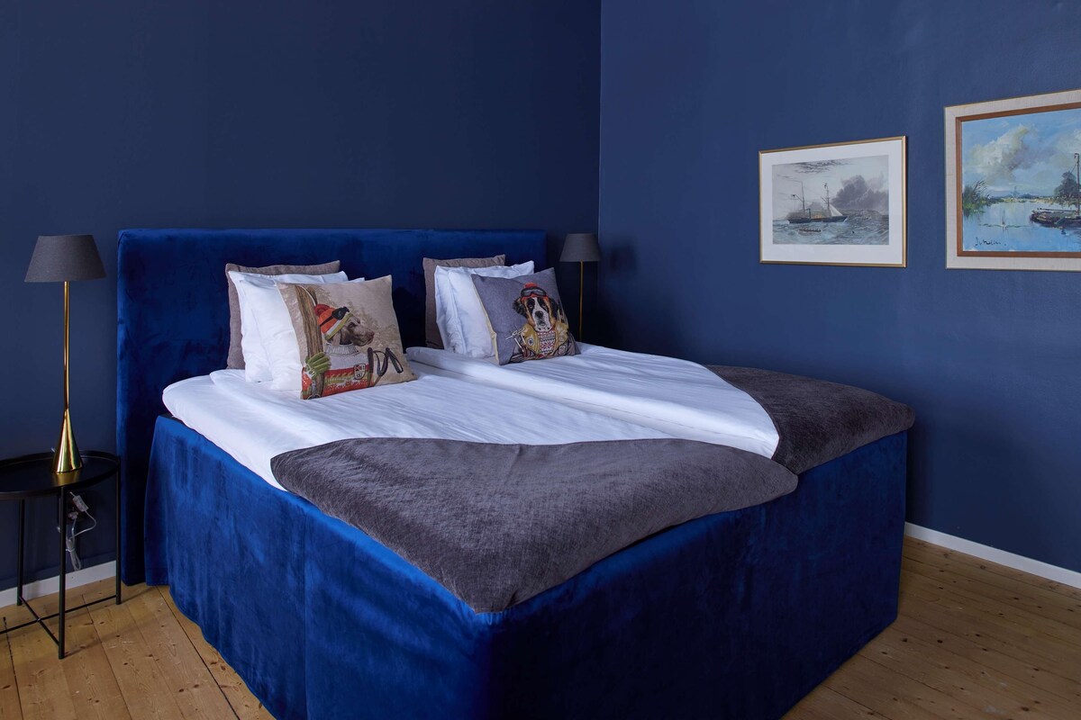 A neatly made bed with a blue headboard, white linens, and a gray blanket. There are two framed artworks on the wall, a lamp on a side table, and a wooden floor.