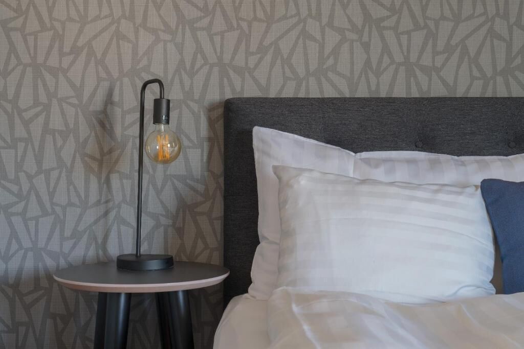 A neatly made bed with white and blue pillows, a round side table with a modern lamp, and a geometric patterned wall.