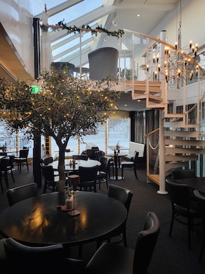 A modern indoor space with a central tree adorned with lights, surrounded by tables and chairs. - Aurum Hotel (Skelleftea)