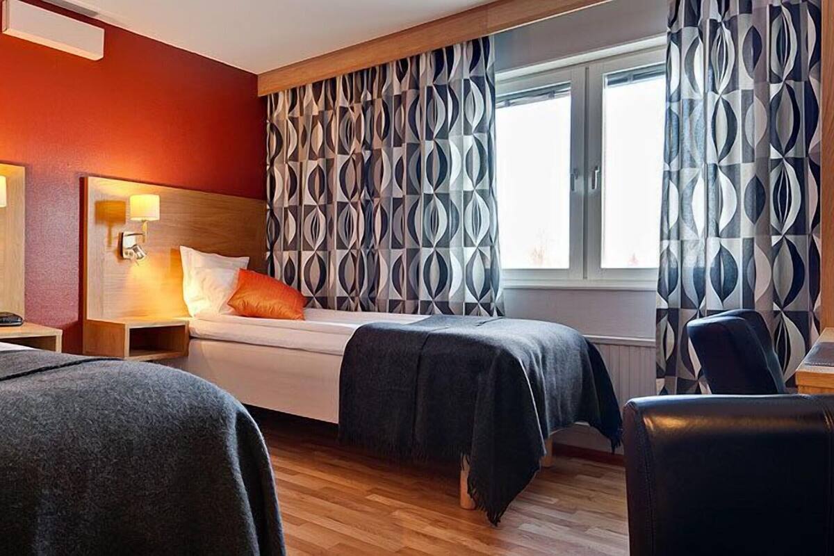 A hotel room with two beds, a patterned wall, a window with curtains, and a wooden headboard.