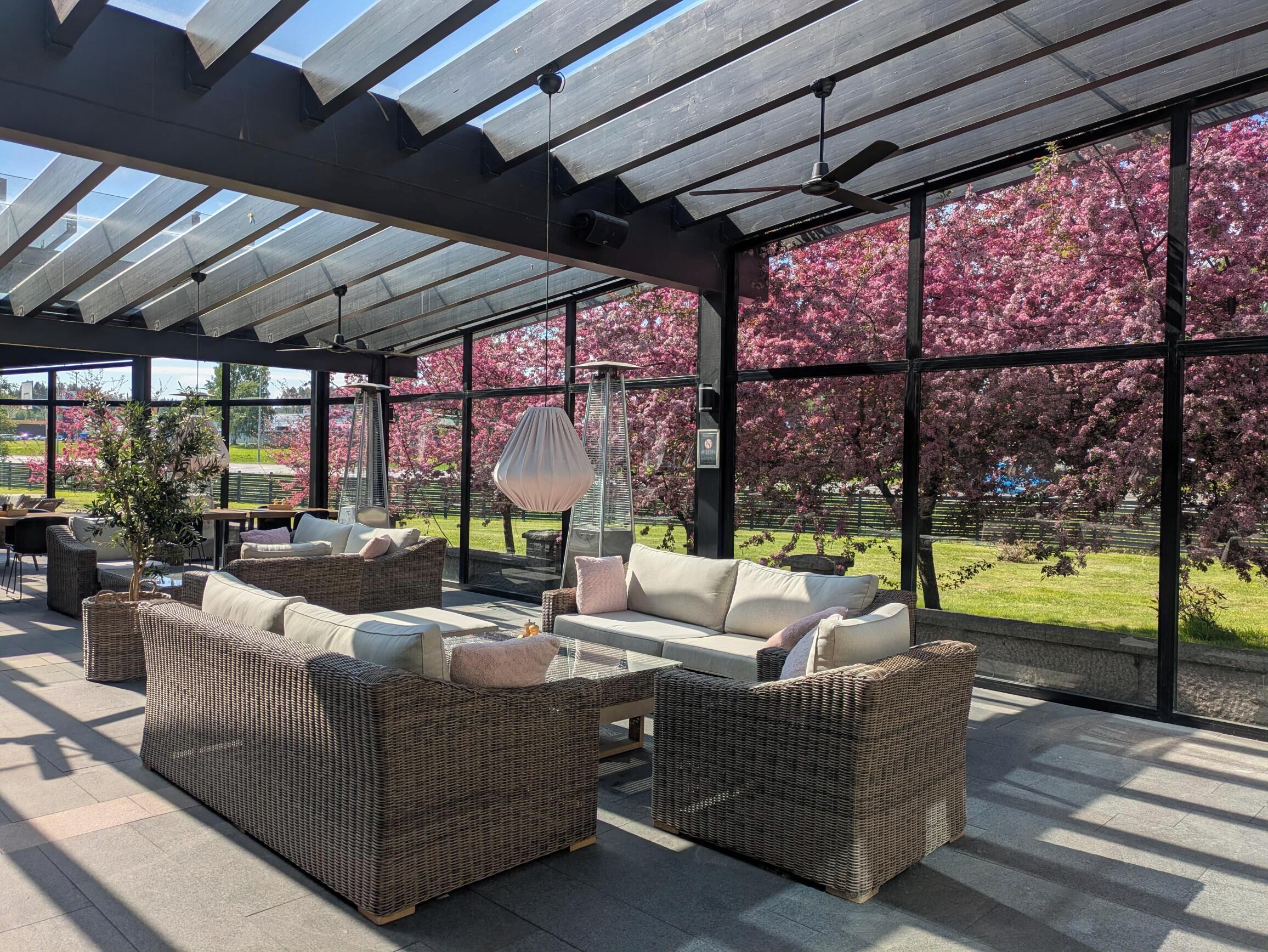 A covered outdoor seating area with wicker furniture and large windows overlooking a garden with pink flowering trees.