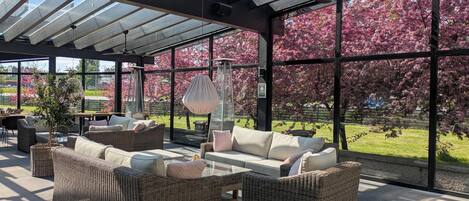 Un espacio al aire libre techado con mobiliario de mimbre y amplias ventanas con vistas a un jardín de árboles de flores rosadas.
