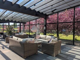 A covered outdoor seating area with wicker furniture and large windows overlooking a garden with pink flowering trees.