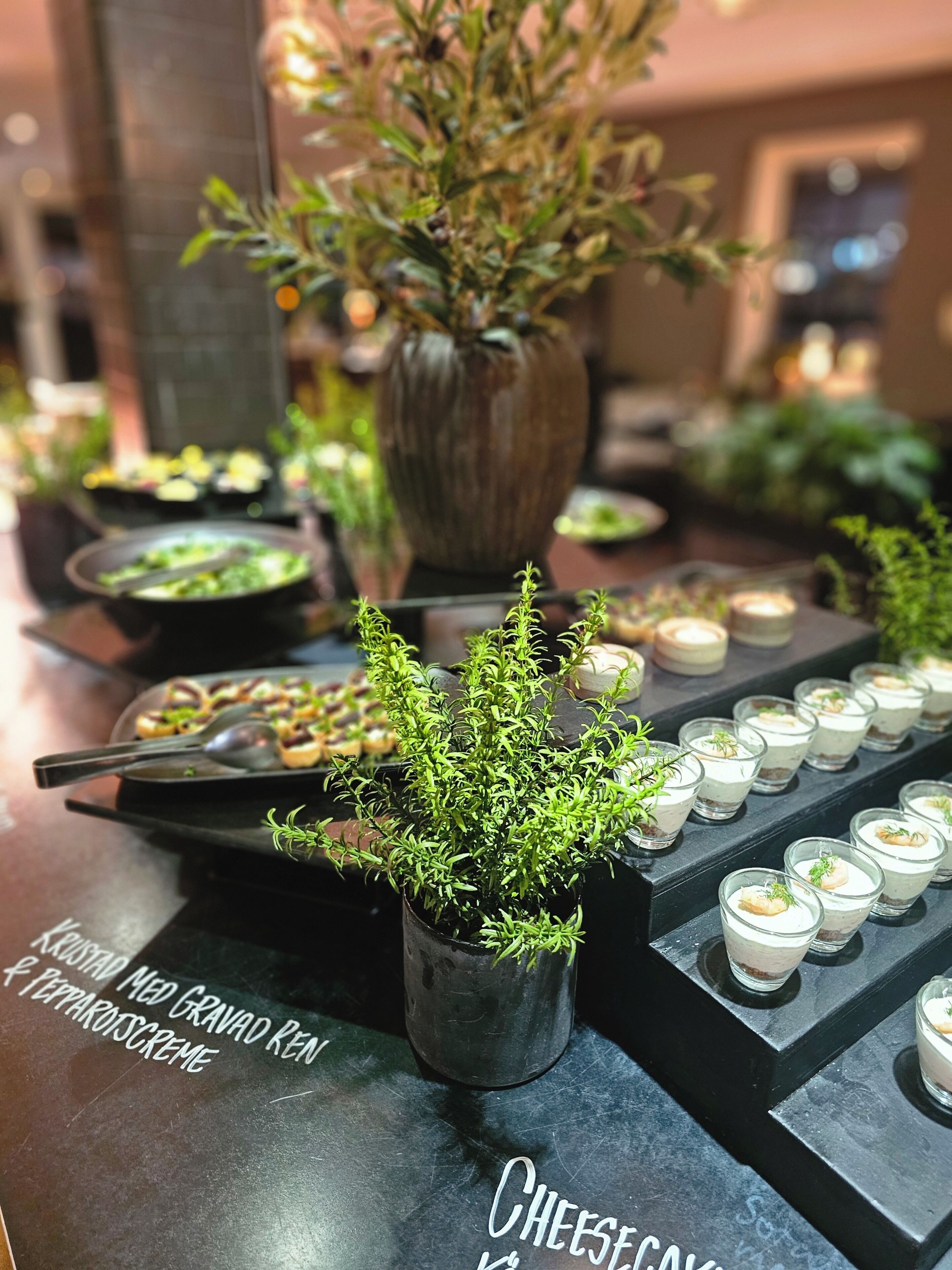 Ein reichhaltiges Buffet mit verschiedenen Speisen, einem kleinen Topfpflanzen und einem Schild, das eine Käseauswahl anzeigt.