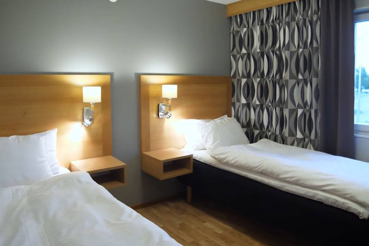 A hotel room with two beds, wooden headboards, bedside tables with lamps, and a patterned curtain.
