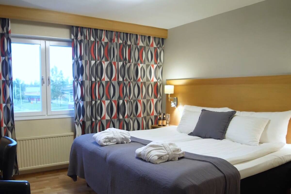 A hotel room with a large bed, a headboard, a window with curtains, a nightstand with a lamp, and a view of the outside.