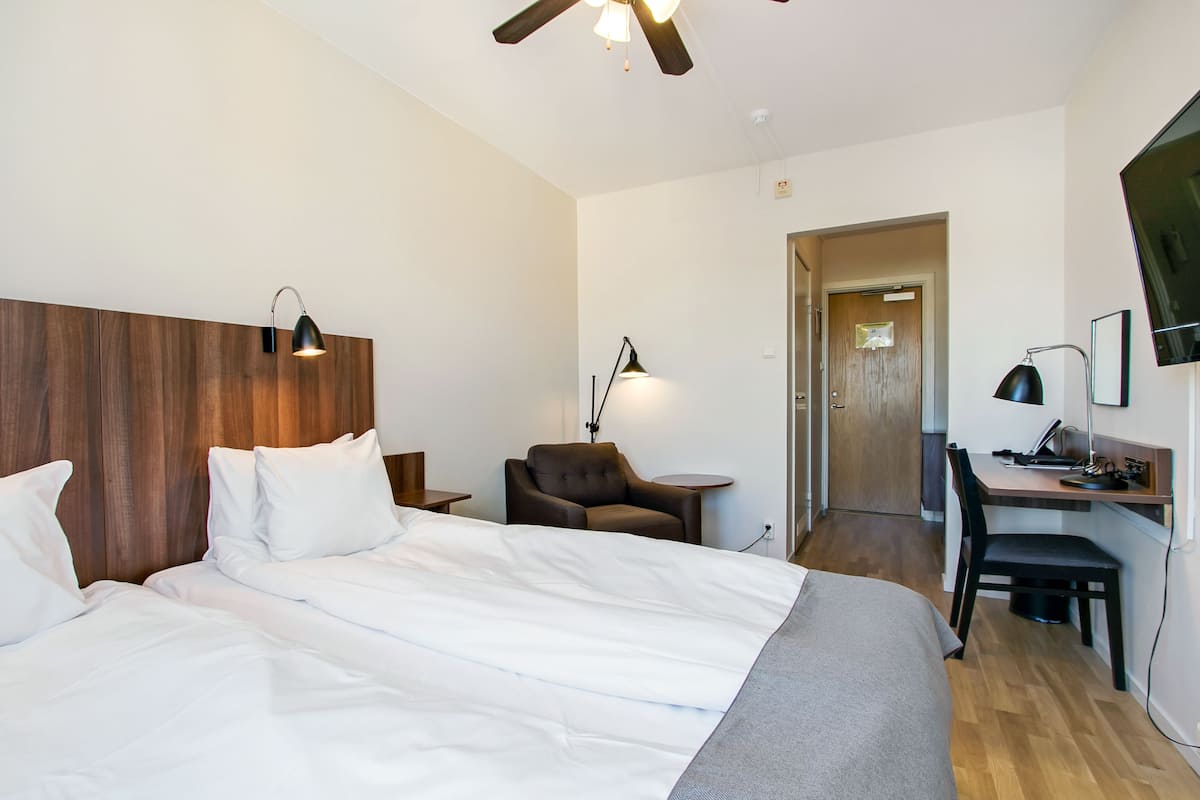 A hotel room with a large bed, a desk with a chair, a ceiling fan, and a TV mounted on the wall.