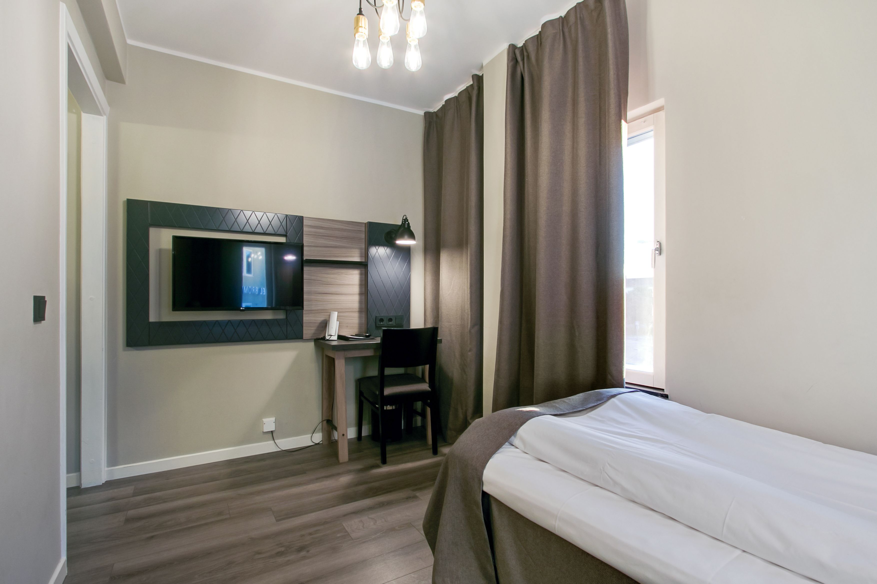 A hotel room with a bed, a desk with a chair, a television, and a window with curtains.