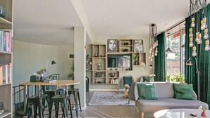 A modern living room with a sofa, coffee table, and bar stools. There is a bookshelf filled with books and decorative items. A television is mounted on the wall, and a window with curtains is visible. - First Hotel Brommaplan (Bromma)