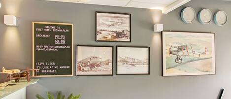 A wall with framed pictures of airplanes, a welcome sign, and clocks.