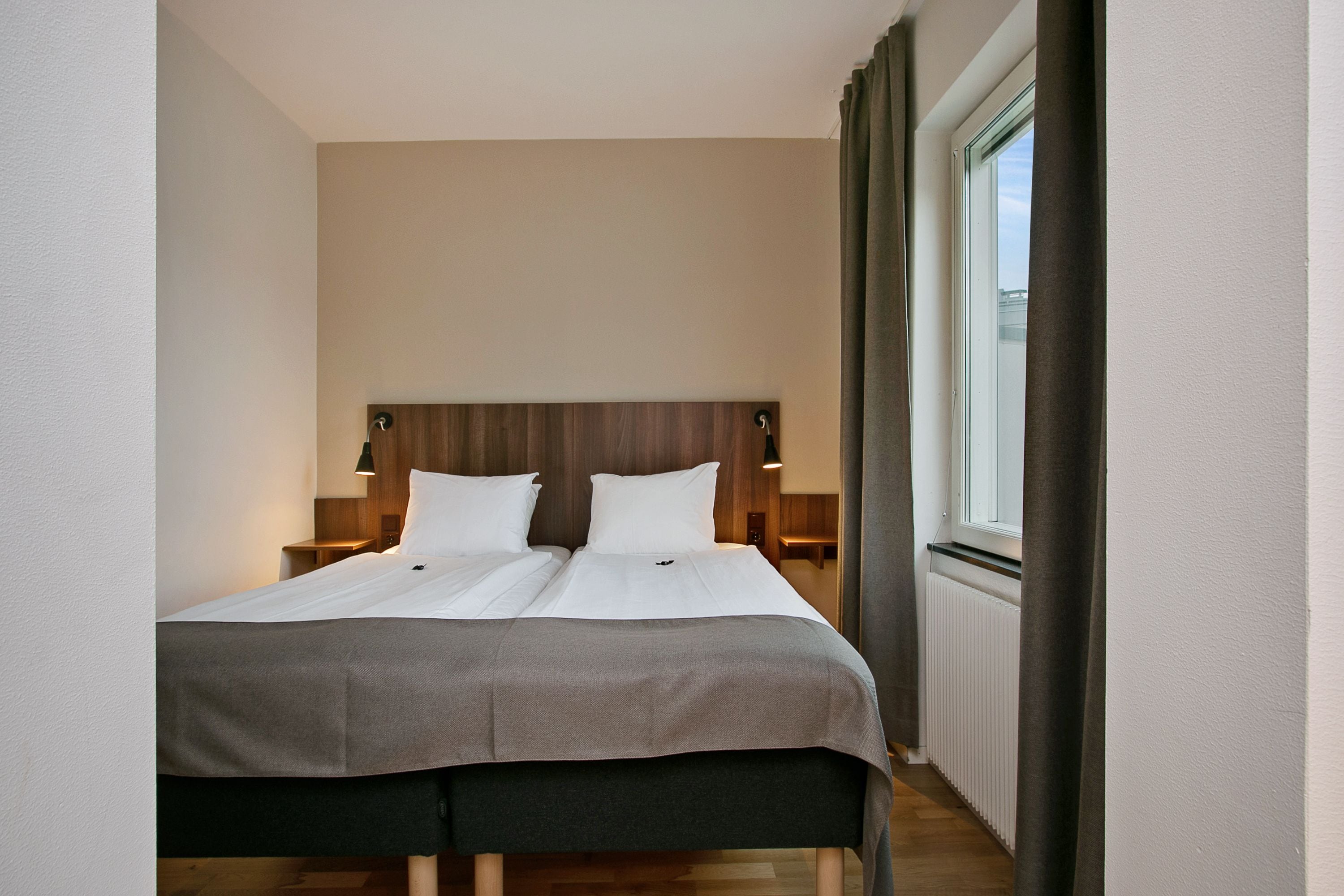 A neatly made twin bed with a wooden headboard, two bedside tables with lamps, and a window with curtains.