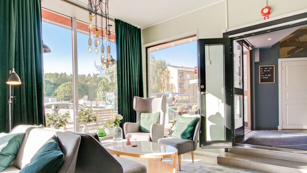 A modern hotel room with a large window, a sofa, a coffee table, and a chair. - First Hotel Brommaplan (Bromma)