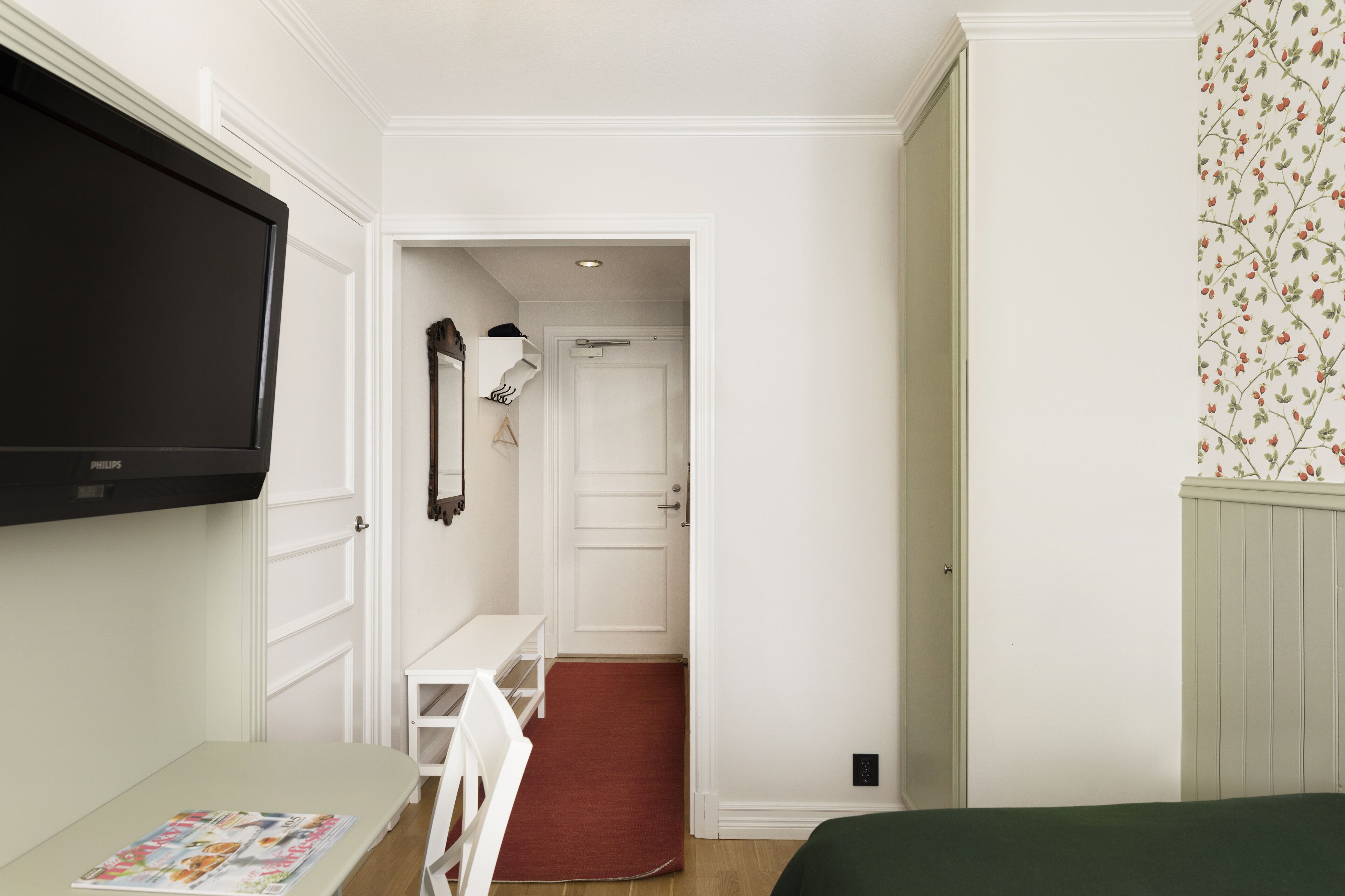 Ein Hotelzimmer mit Fernseher, Schreibtisch mit Zeitschrift, rotem Teppich und Bett.