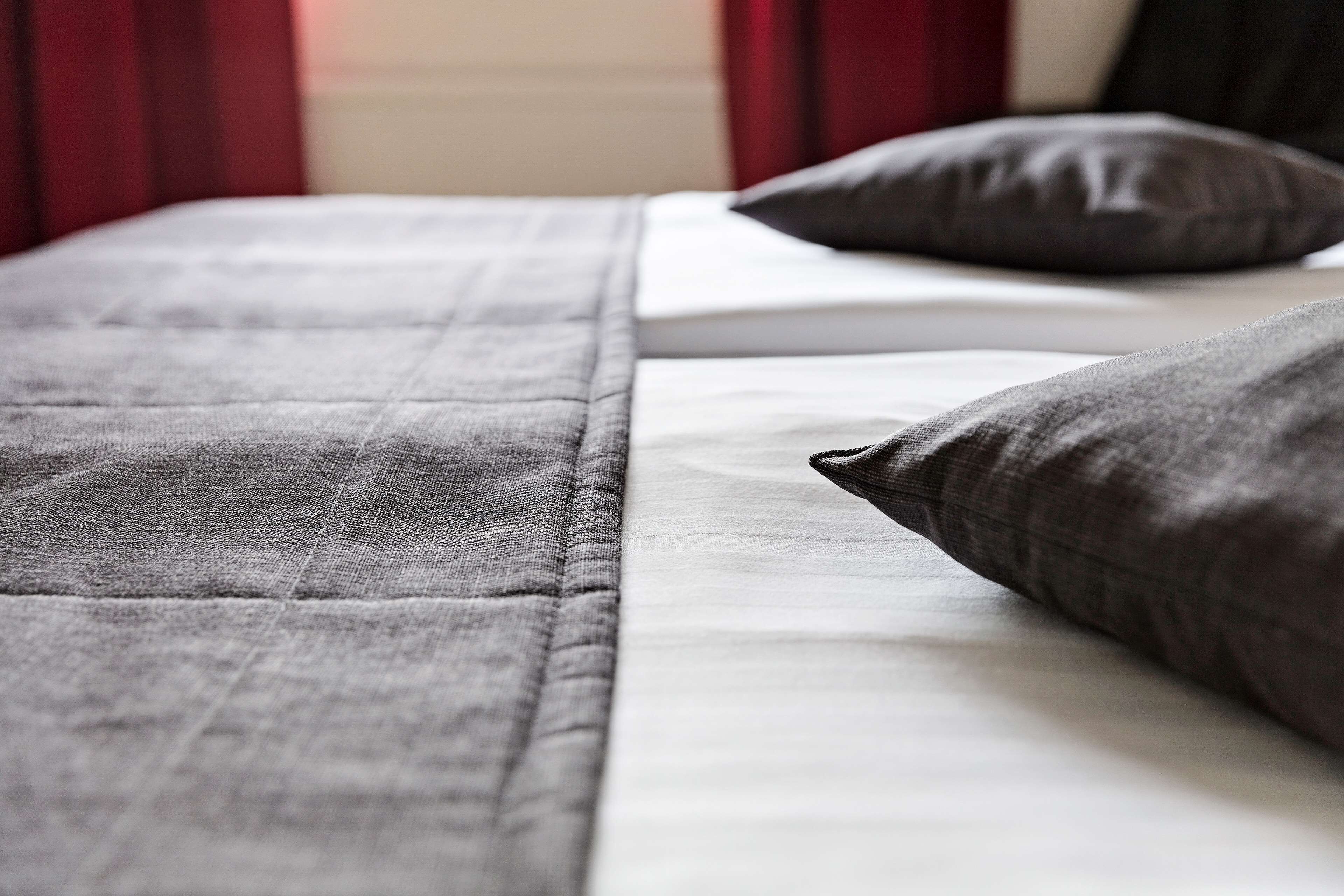 A bed with grey bedding and pillows, a white mattress, and red curtains in the background.