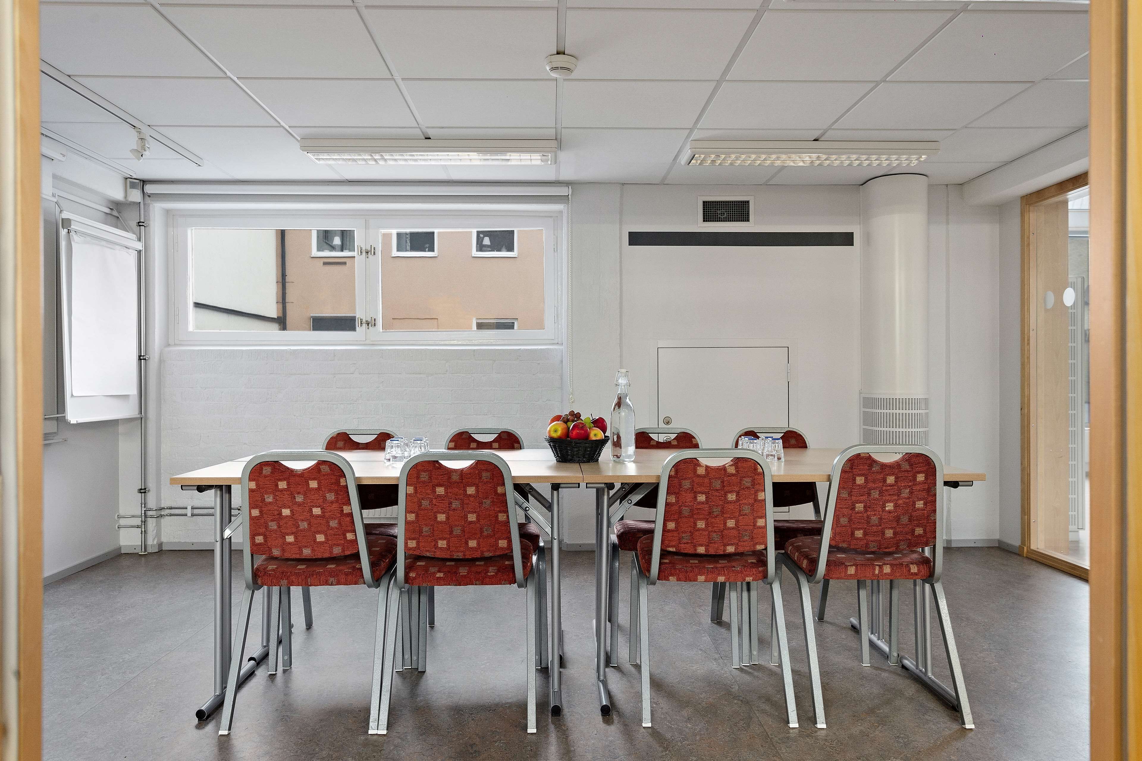 A conference room with a long table, six red patterned chairs, a fruit basket, and a water bottle.