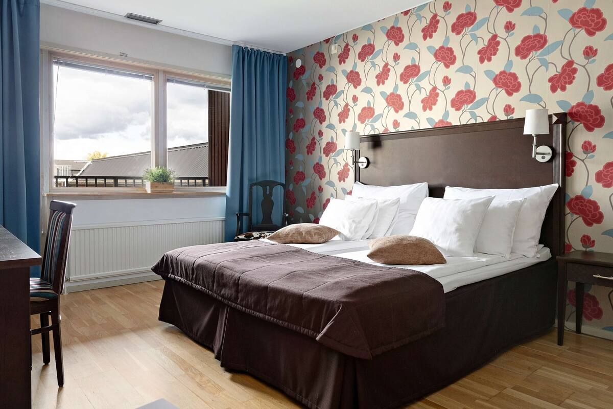 A hotel room with a large bed, a desk, and a chair. There is a window with curtains, a wall with floral wallpaper, and a nightstand with a lamp.