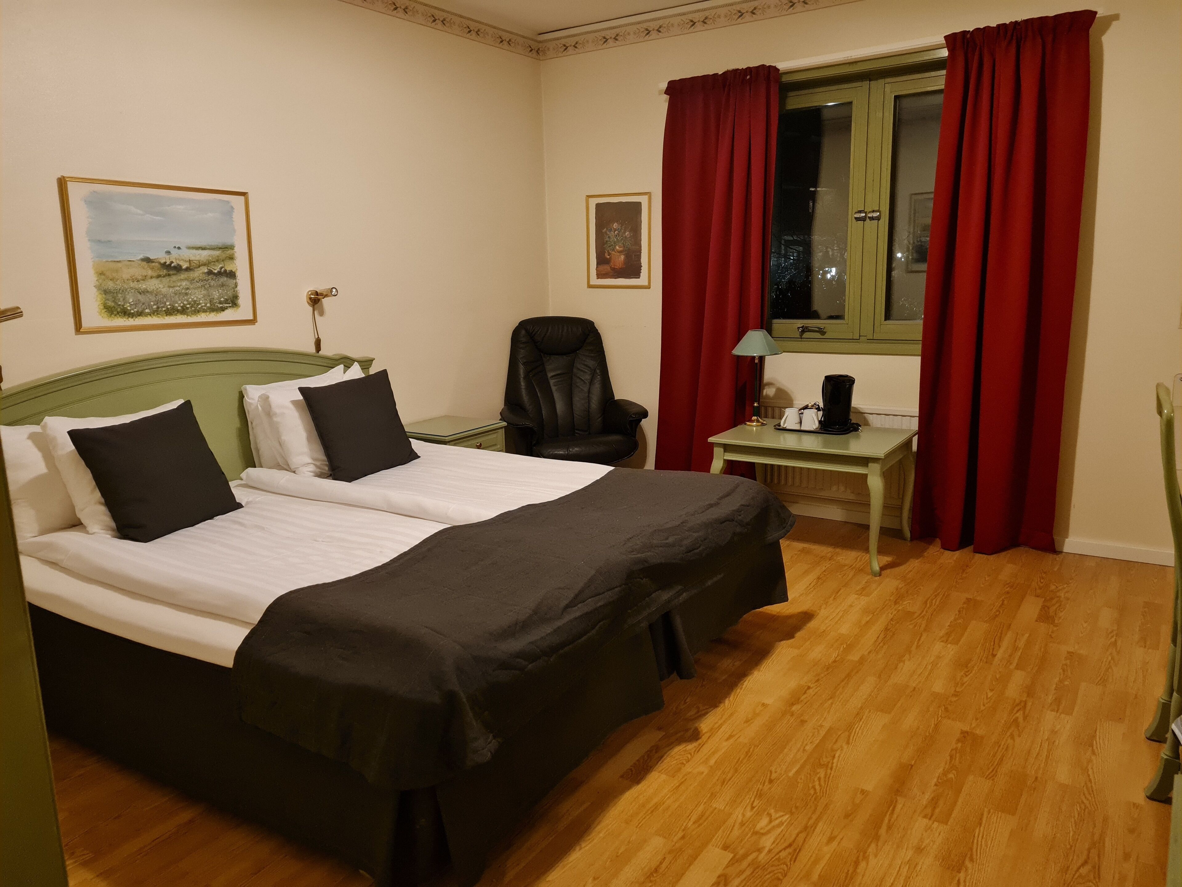 A double bed with white and black bedding, a green headboard, a small table with a lamp, a chair, and a window with red curtains.