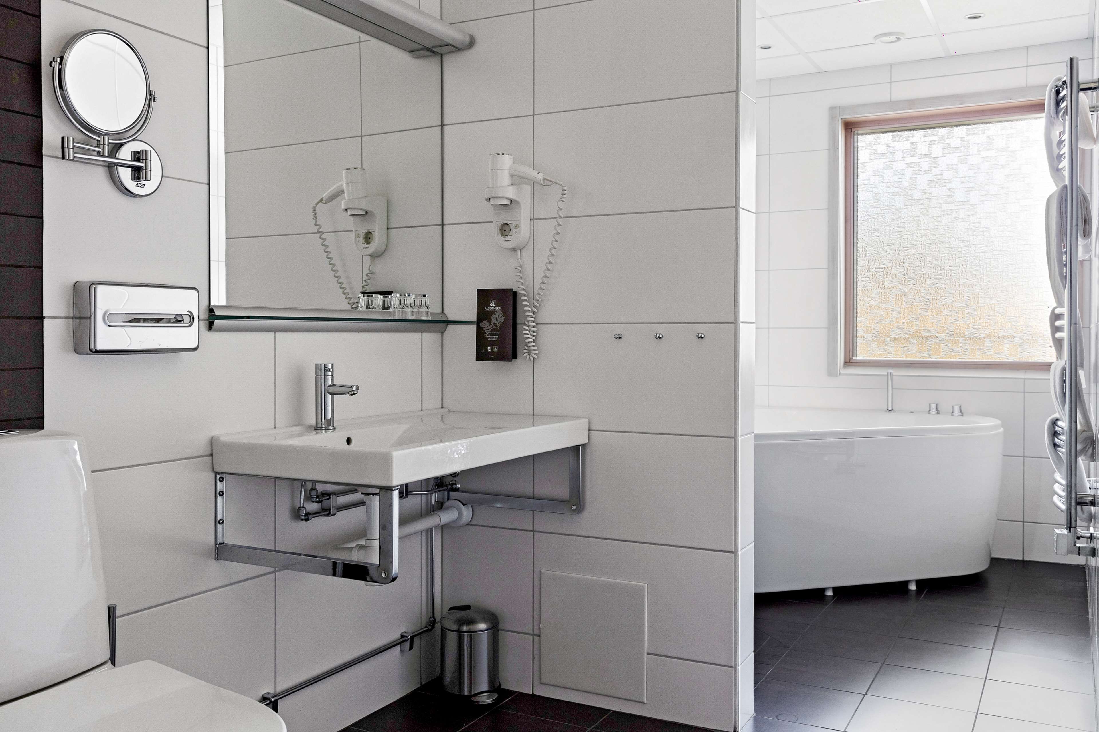 Un baño moderno con lavamanos, espejo y retrete blancos. Cuenta con cabina de ducha con puerta de cristal y ventana que proporciona luz natural.