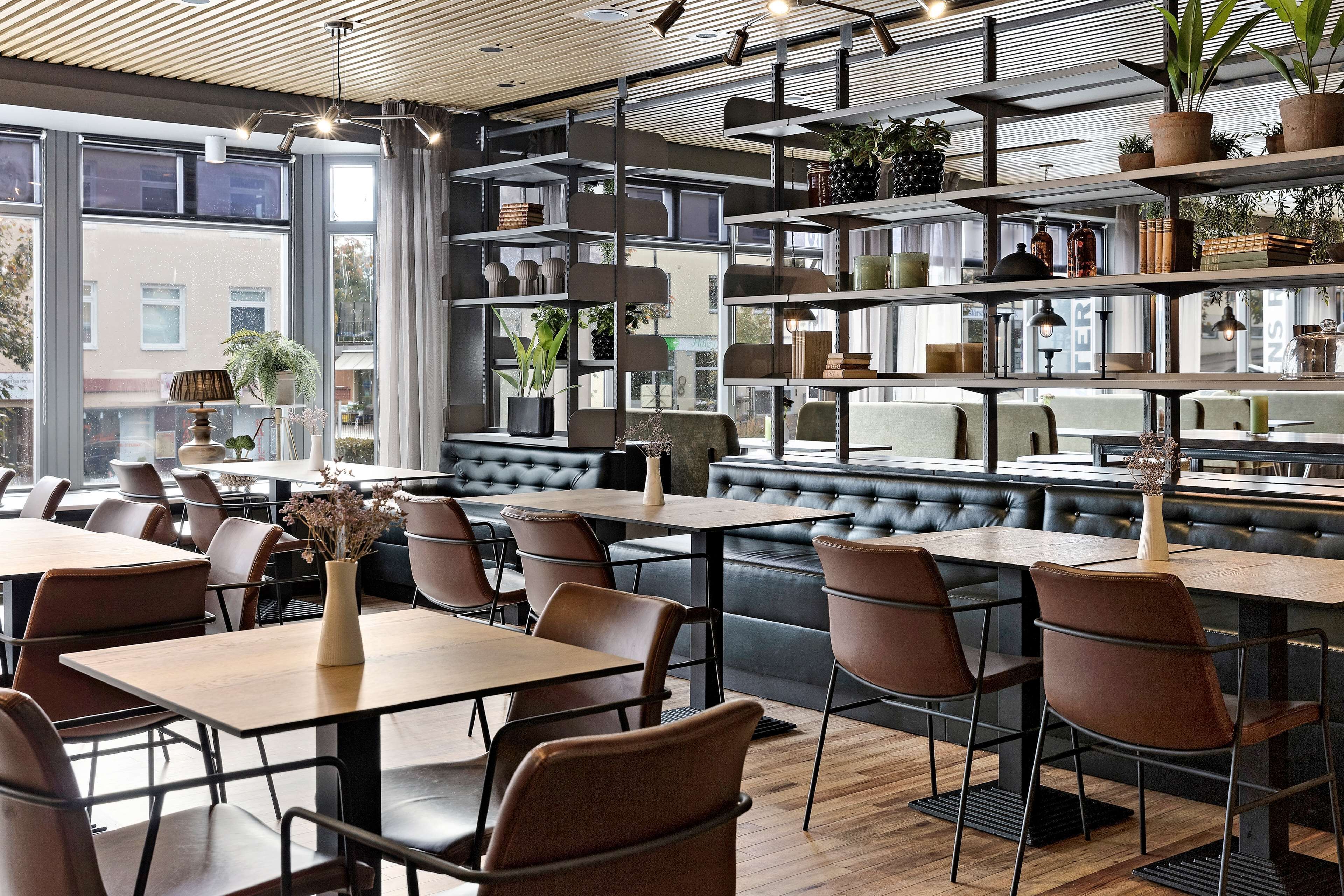 A modern restaurant with wooden tables and chairs, a bar area with a high counter, and shelves with plants and books.