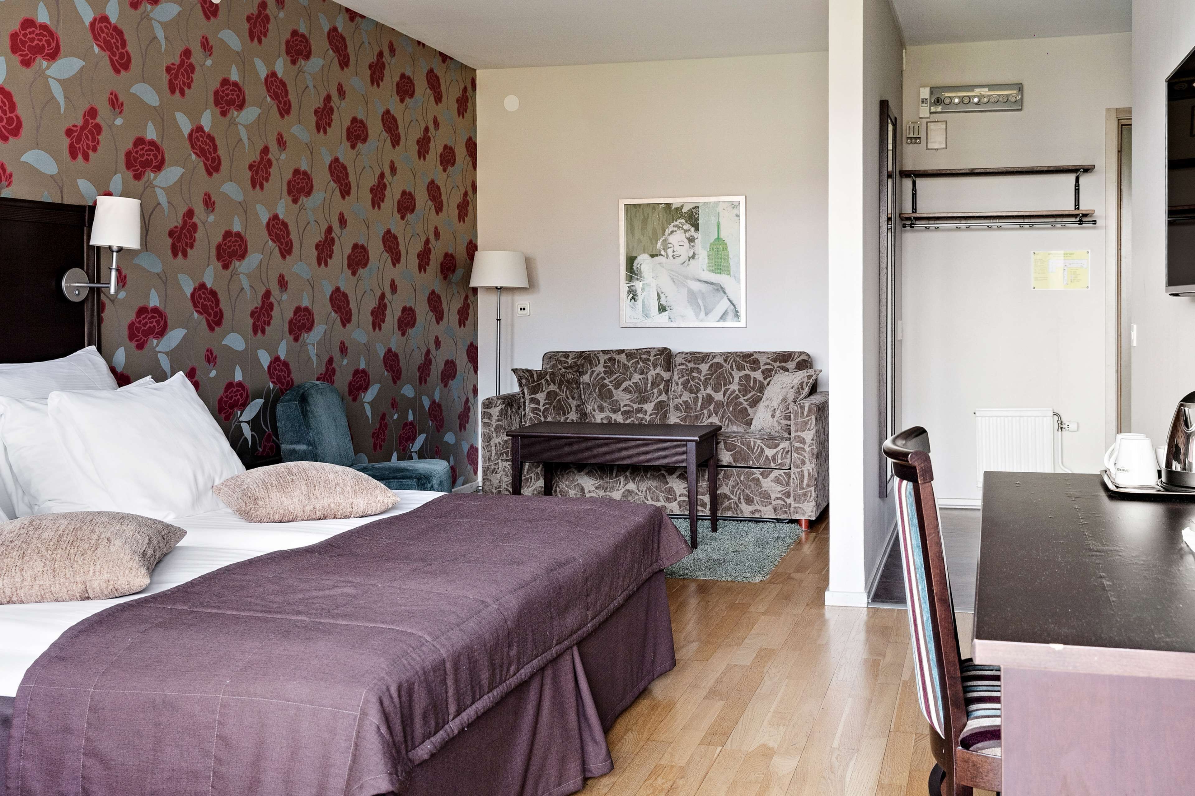 A hotel room with a bed, a sofa, a small table, and a chair. There is a painting on the wall and a radiator above the sofa.