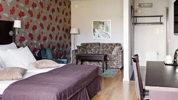 A hotel room with a bed, a sofa, a small table, and a chair. There is a painting on the wall and a radiator above the sofa.