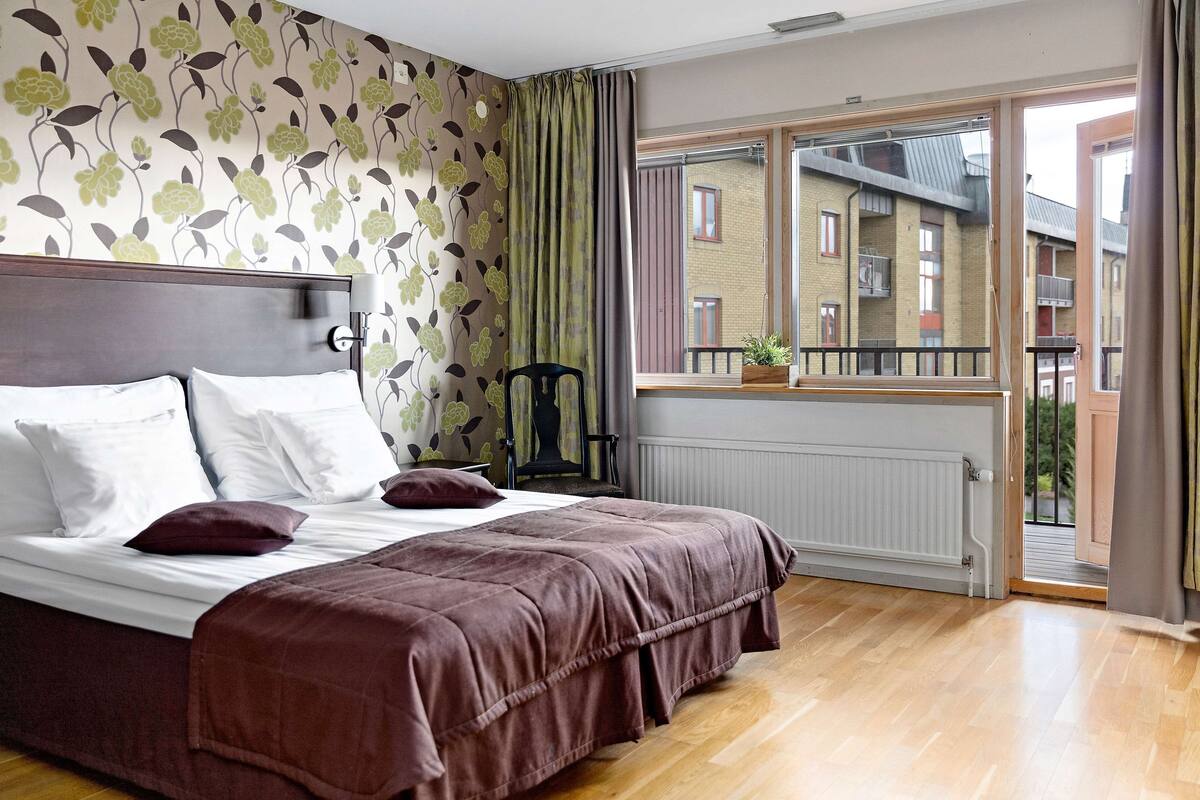 A bedroom with a large bed, a radiator, a window with a view of buildings, and a curtain.