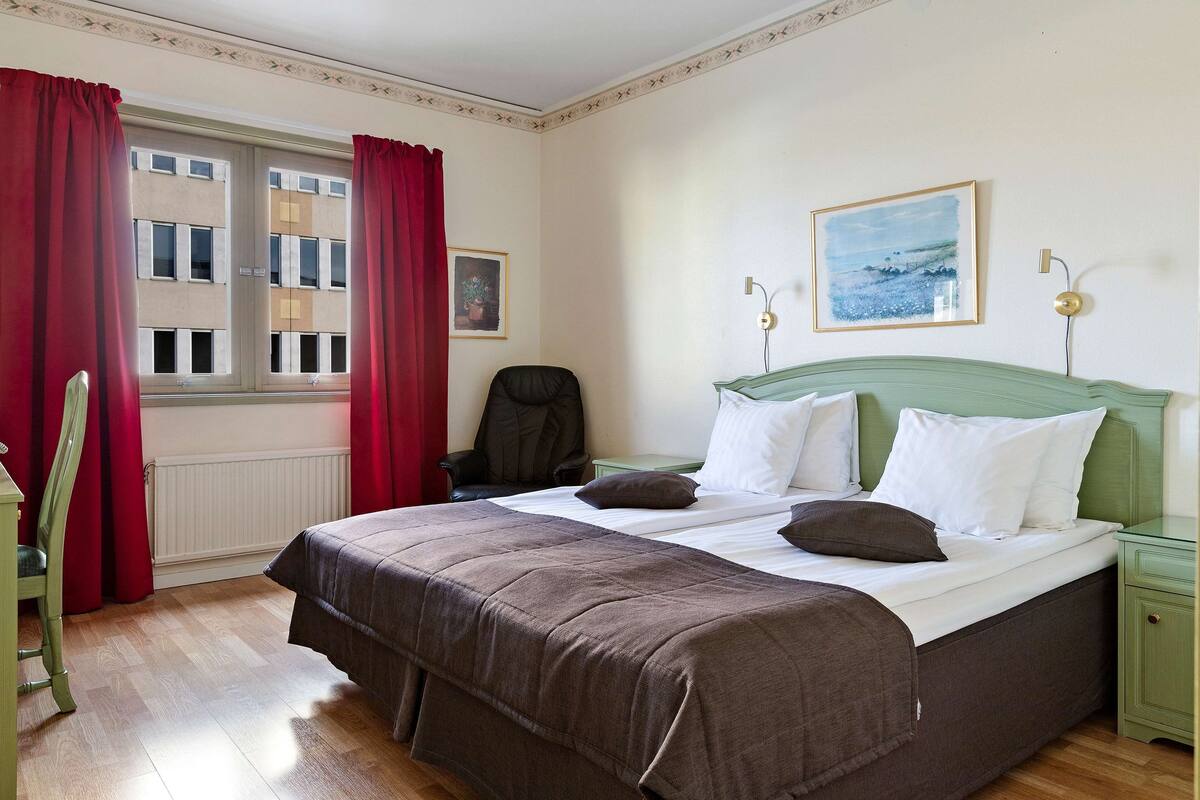 A bedroom with a large bed, a chair, a desk, and a window with red curtains.