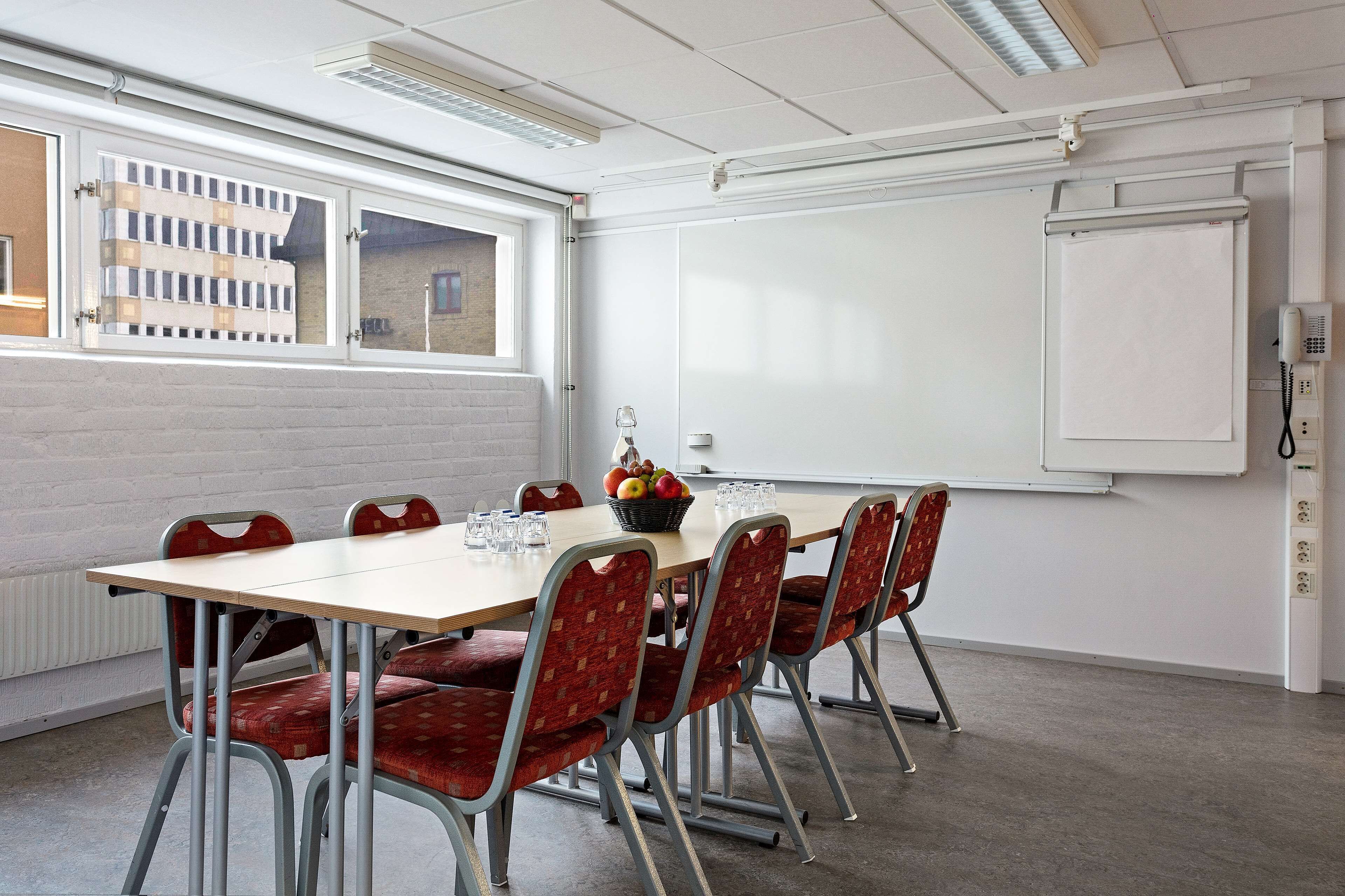A conference room with a long table, red upholstered chairs, a whiteboard, and a window with a view of buildings.
