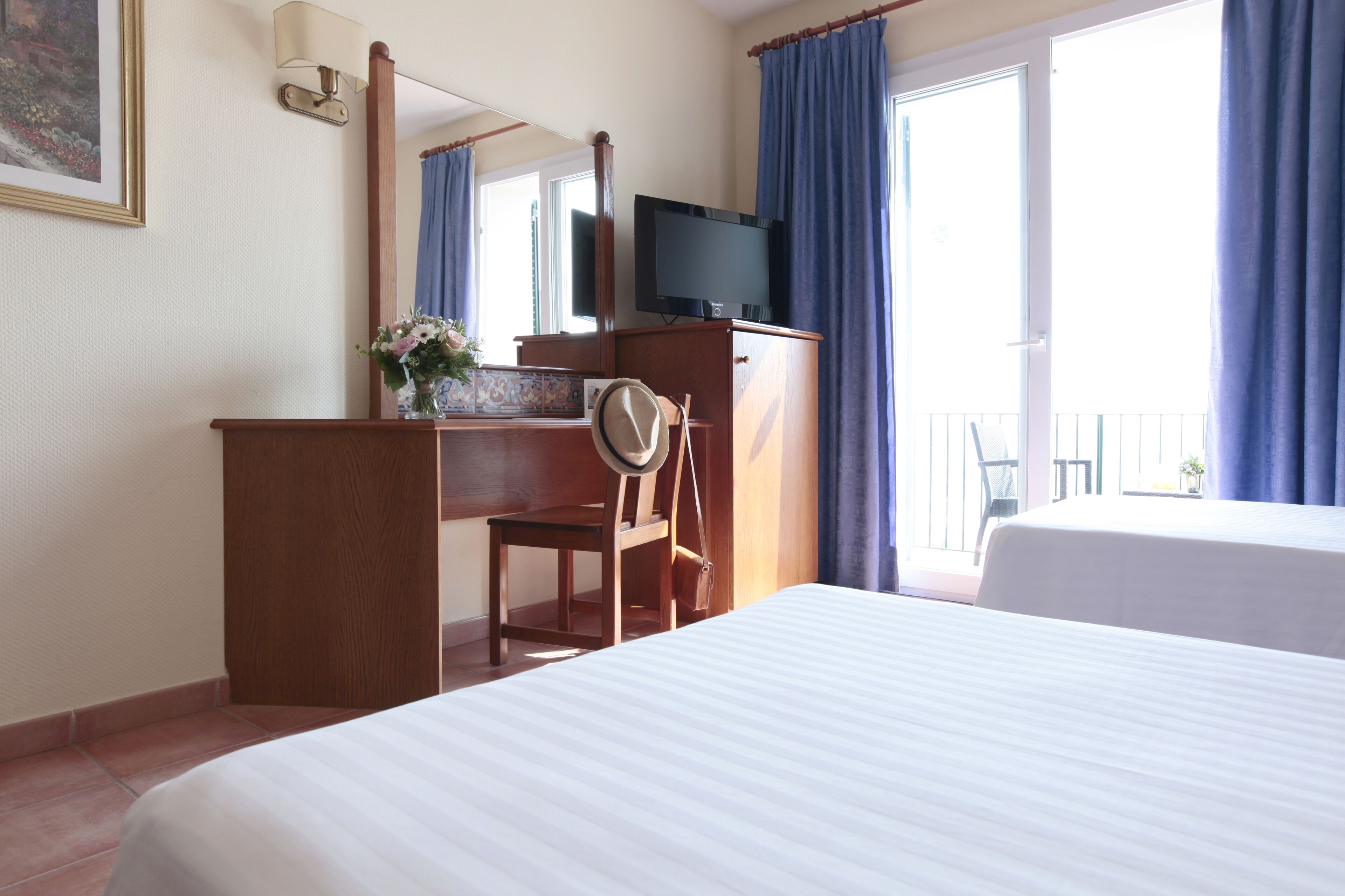 A hotel room with a bed, a desk with a television, a chair, and a balcony with curtains.