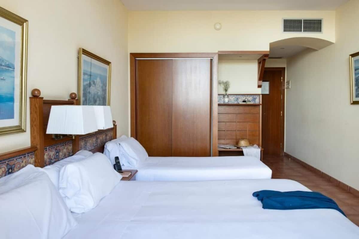 A hotel room with two beds, a wooden wardrobe, a bathroom with a sink and mirror, and a small balcony.