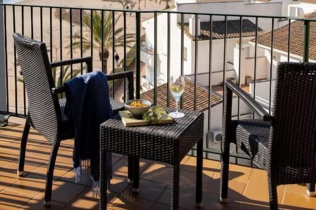 A balcony with wicker chairs and a table, overlooking a beach and buildings.