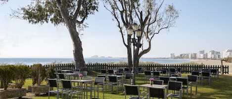 An outdoor seating area with tables and chairs arranged under trees, overlooking a beach and ocean.