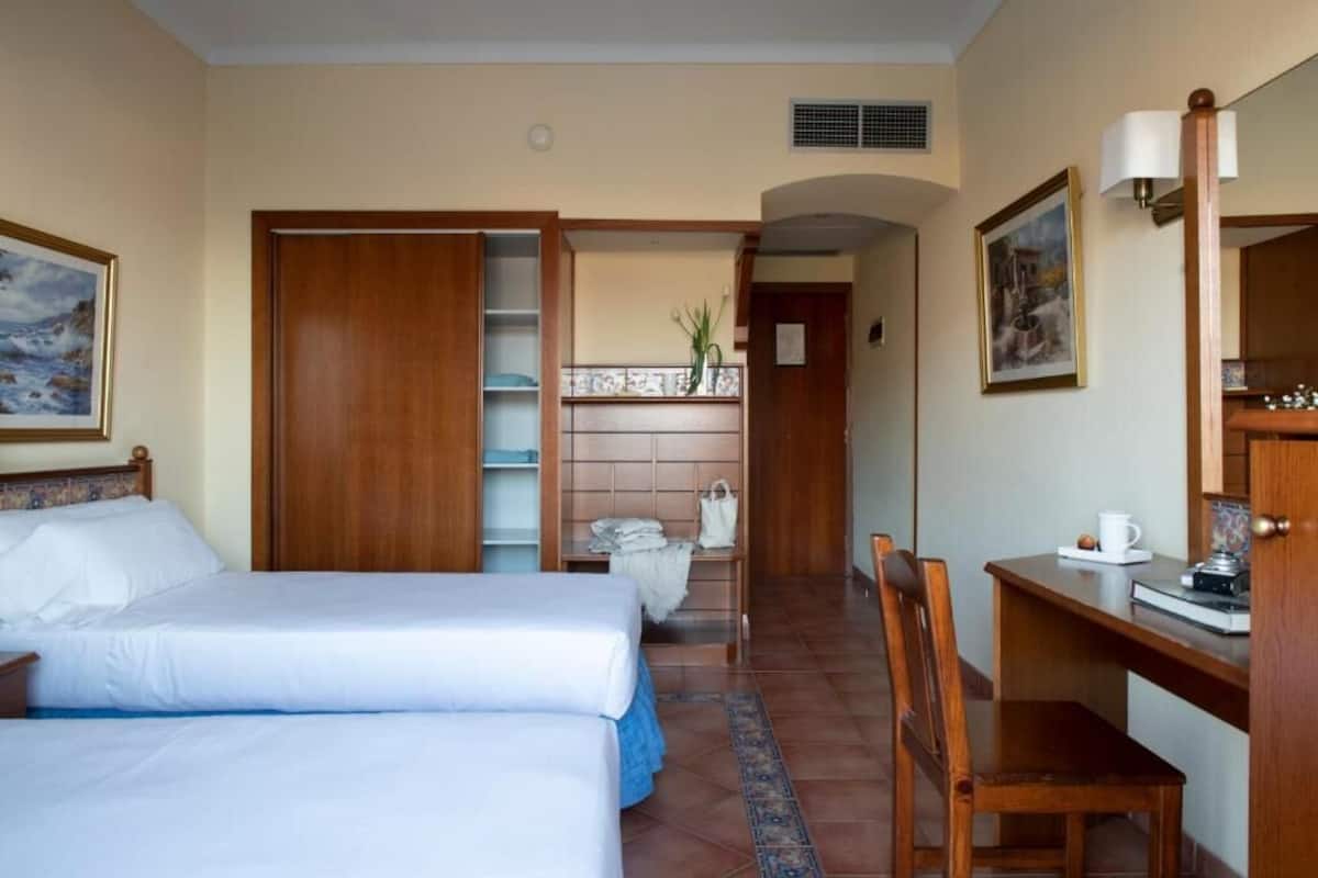 A hotel room with two beds, a desk, a chair, a wardrobe, and a bathroom visible through an open door.