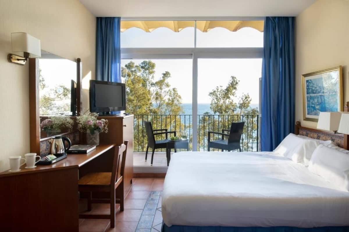A hotel room with a bed, desk, chair, TV, and balcony with a view.