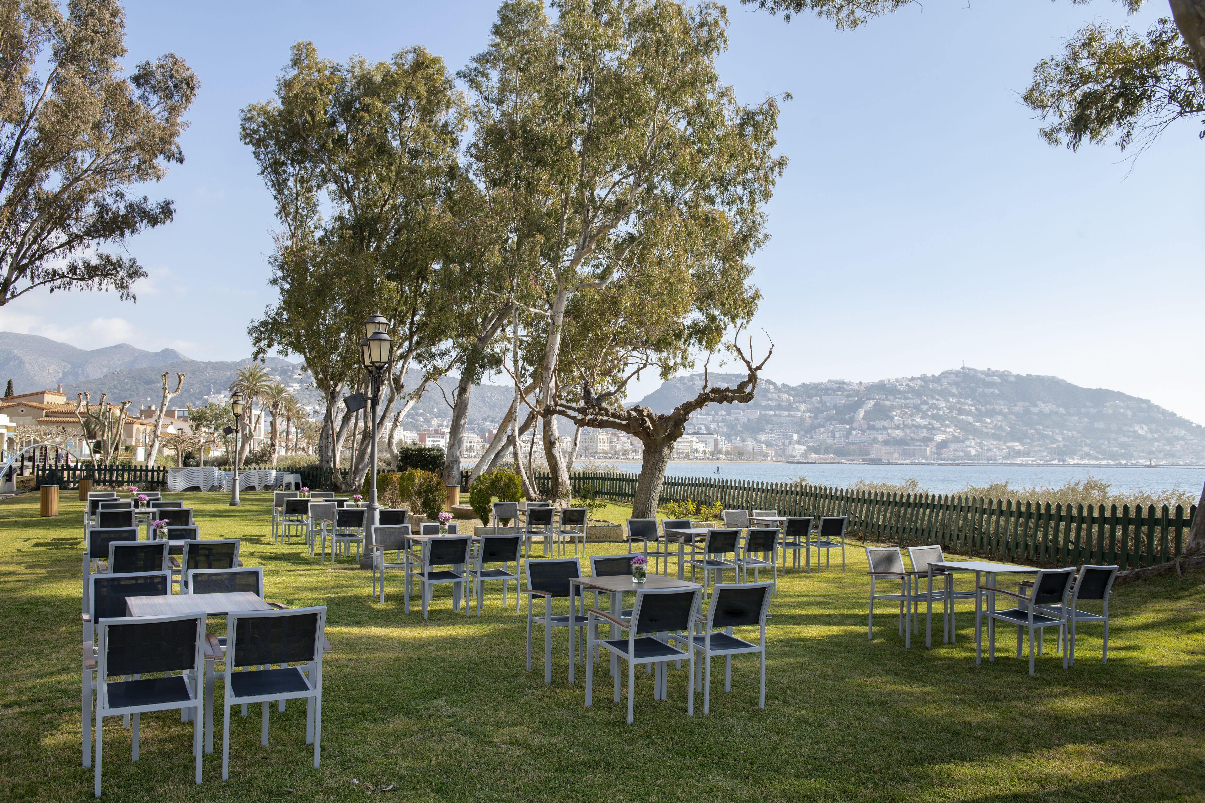 Un espace extérieur aménagé avec des tables et des chaises disposées sur une pelouse, offrant une vue sur une étendue d’eau et un paysage montagneux.