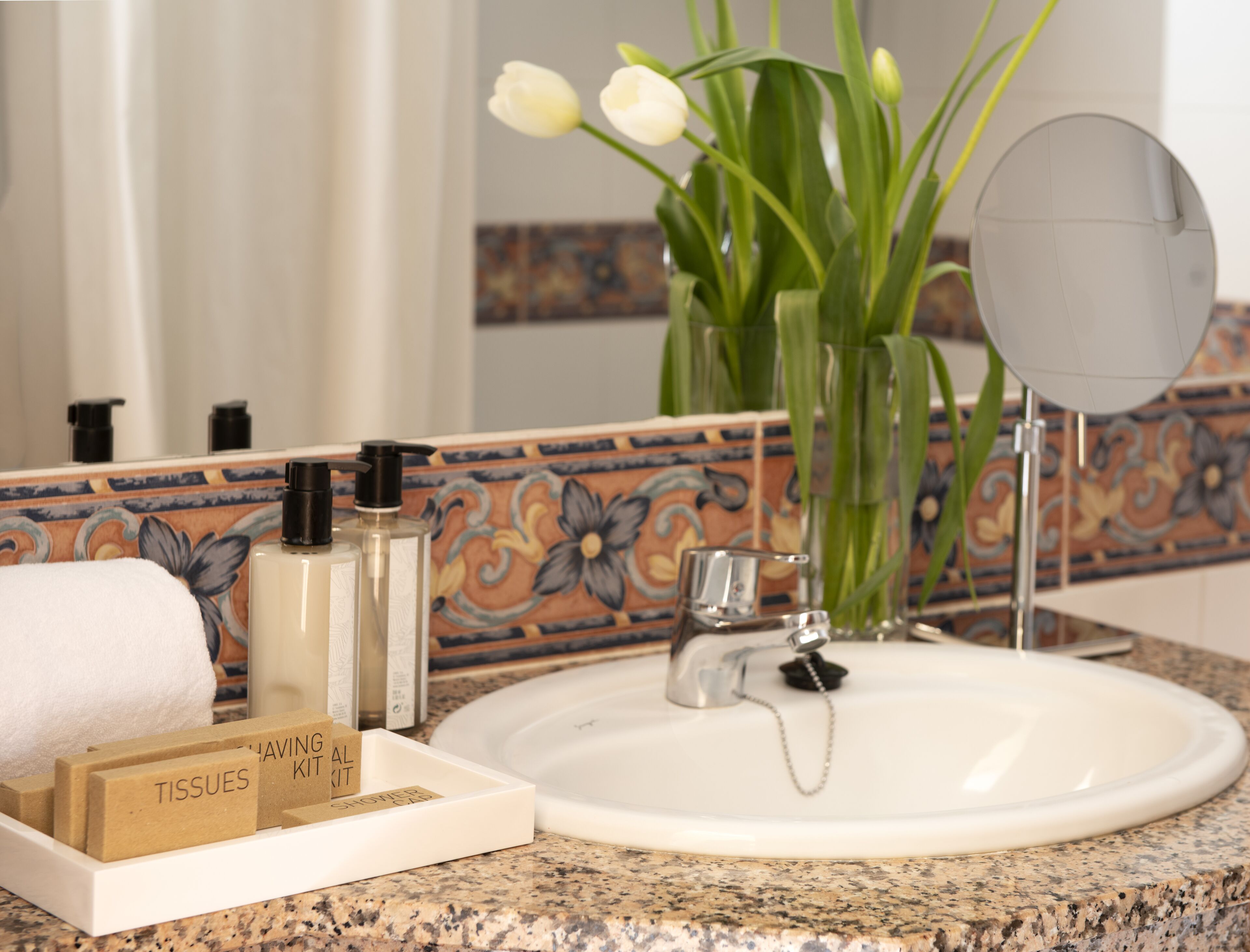 Un baño con un lavabo blanco, un espejo, un florero con flores blancas y artículos de aseo en la encimera.