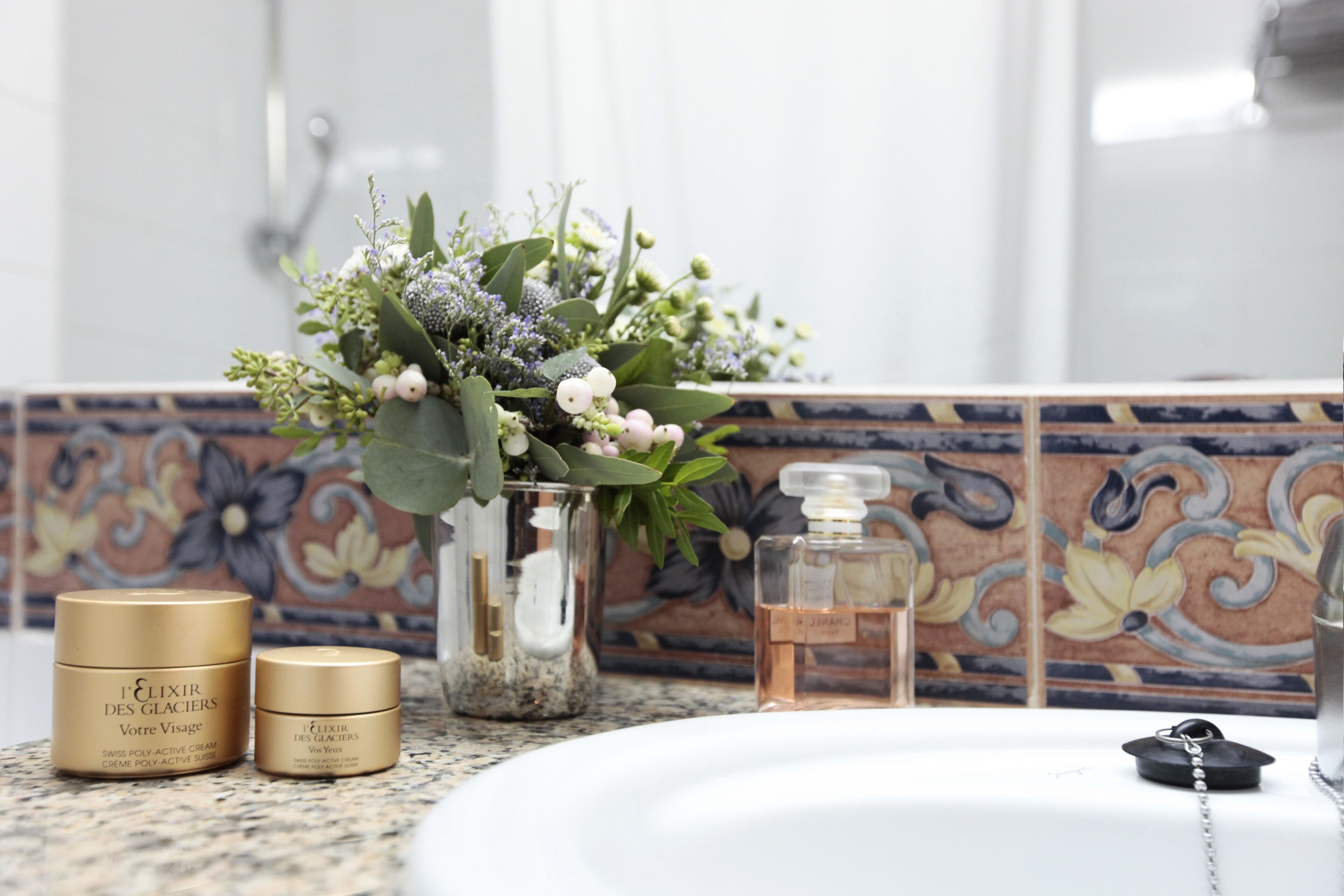 A bathroom vanity with a white sink, a glass vase with flowers, and two gold containers of 'Elixir des Glaciers' cream.