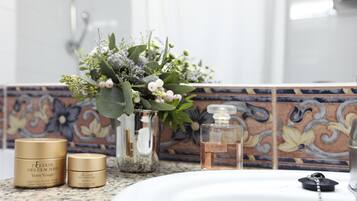 A bathroom vanity with a white sink, a glass vase with flowers, and two gold containers of 'Elixir des Glaciers' cream.