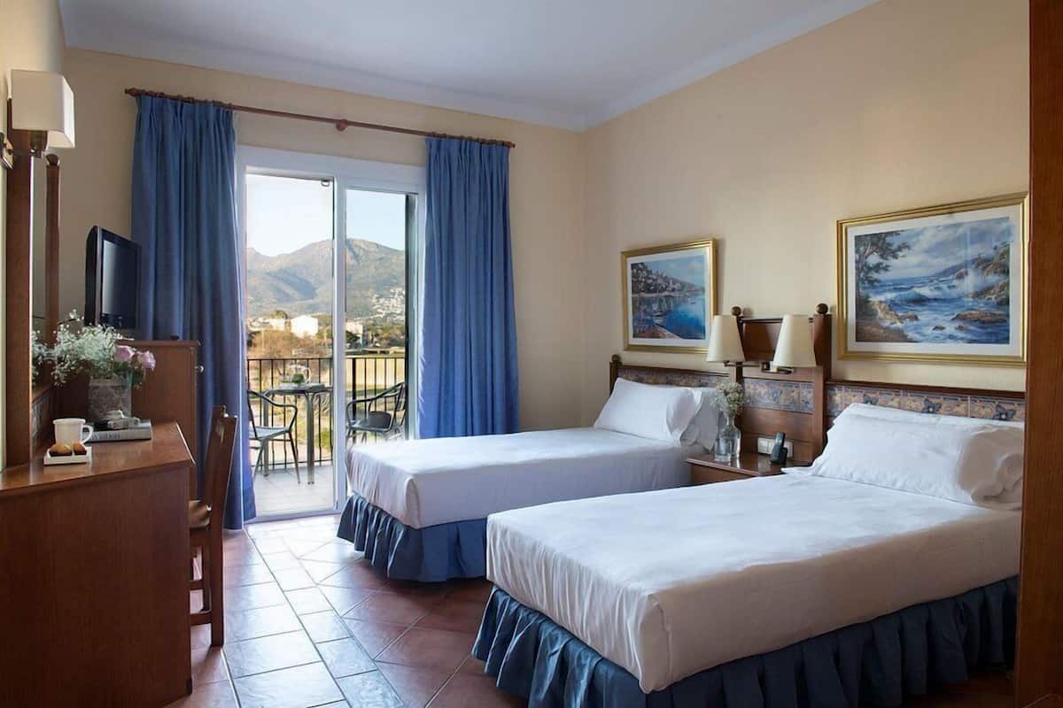 A hotel room with two beds, a desk, a chair, a TV, and a balcony with a view of the mountains.